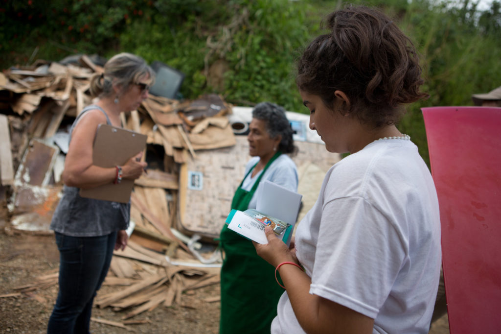 Jumileysie de Jesus de Jesus, 15, looks at photos of her family's damaged home as Silvia XX, left, a volunteer with the local organization Casa Pueblo, which is partnering with LWR in Puerto Rico, speaks to her grandmother Severina de Jesus Hernandez, 74, in front of a pile of ruined belongings dragged from the house after Hurricane Maria inundated the dwelling with water six weeks ago, in the town of Vegas Arriba, in the municipality of Adjuntas, Puerto Rico, November 2, 2017. (photo by Allison Shelley for Lutheran World Relief)